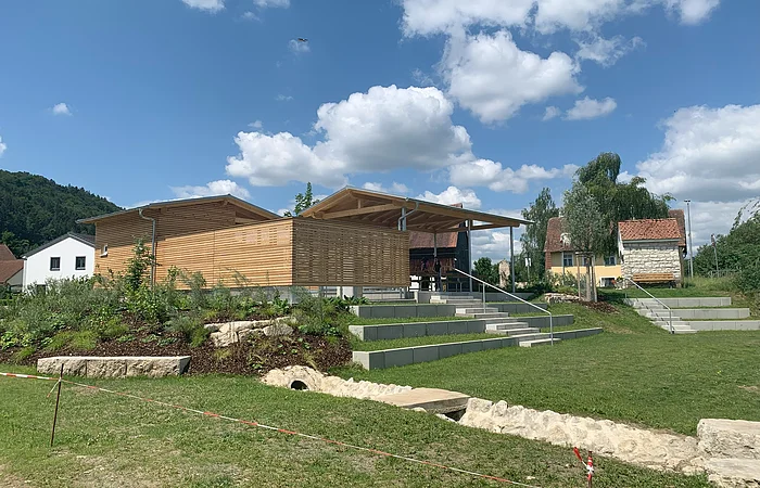 Ein modernes Gebäude mit Holzverkleidung und überdachter Terrasse, umgeben von Grünflächen und Treppenstufen.