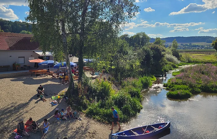 Menschen entspannen auf einer Wiese neben einem kleinen Fluss, mit einem Kanu im Vordergrund und einem Gebäude im Hintergrund.