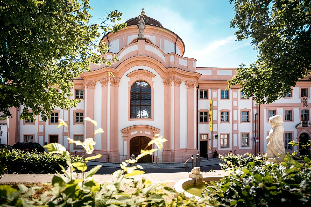 Barockgebäude mit großer Fensterfront, Statue auf dem Dach und Springbrunnen im Vordergrund bei Sonnenschein