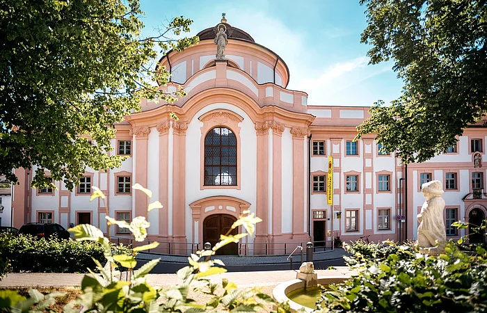 Barockgebäude mit großer Fensterfront, Statue auf dem Dach und Springbrunnen im Vordergrund bei Sonnenschein