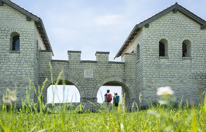 Zwei Personen mit Rucksäcken stehen unter einem steinernen Torbogen einer historischen Anlage bei sonnigem Himmel.