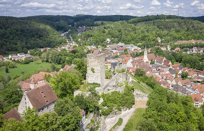 Luftaufnahme einer Burg mit Turm und umliegendem Dorf in einer grünen Hügellandschaft unter bewölktem Himmel.