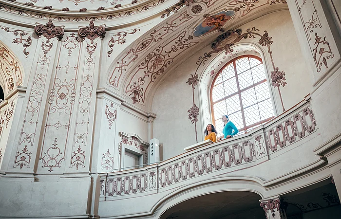 Zwei Personen stehen auf einem verzierten Balkon in einem historischen, mit Fresken geschmückten Innenraum.