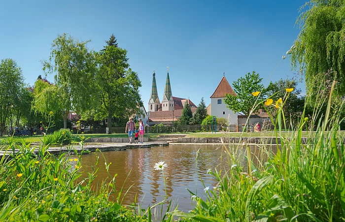 Park mit Teich, Wiese, Bäumen und Menschen vor historischen Gebäuden mit zwei Türmen bei blauem Himmel