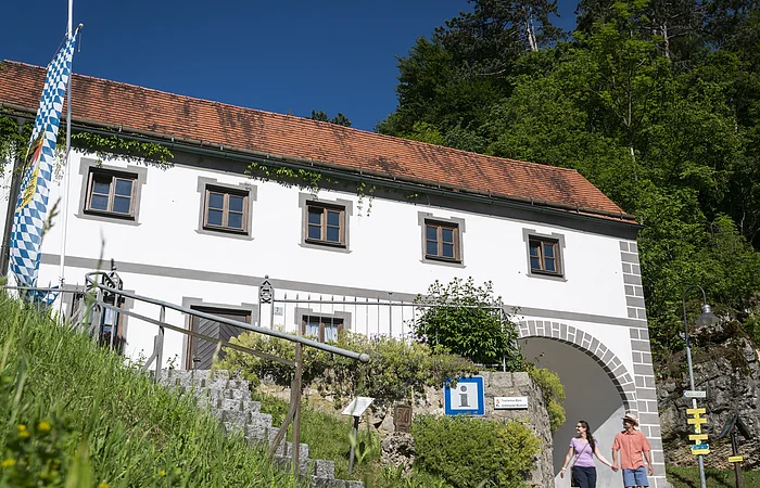 Weißes Gebäude mit rotem Ziegeldach, bayerischer Flagge und zwei Personen vor einem Torbogen bei Sonnenschein