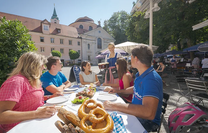 Fünf Personen sitzen an einem Tisch im Freien, eine Kellnerin bringt Bier, Brezeln und Brot liegen auf dem Tisch.