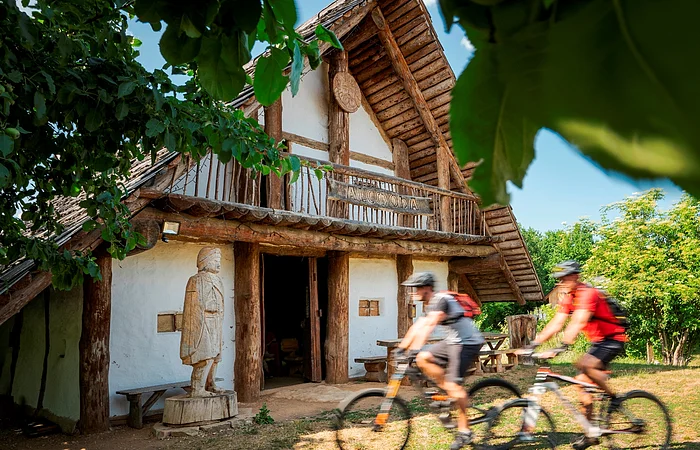 Holzhaus mit Statue davor und zwei Radfahrern, umgeben von Bäumen bei sonnigem Wetter