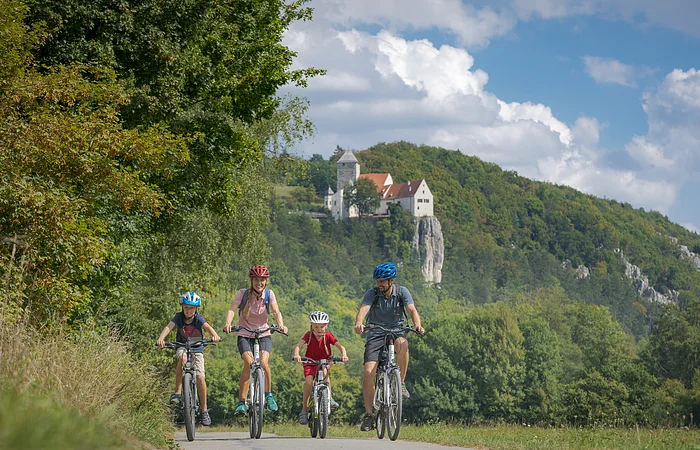 Familie mit zwei Kindern fährt auf Fahrrädern auf einem Weg vor bewaldetem Hügel mit Burg im Hintergrund.