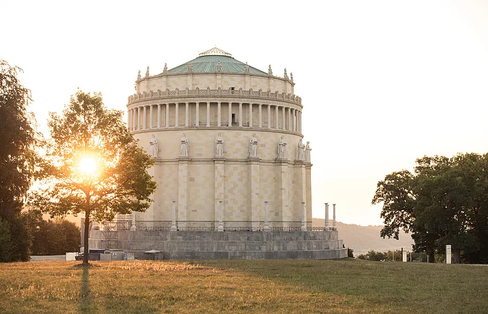 Rundes historisches Gebäude mit Statuen auf dem Sims, Sonne hinter Baum im Park bei Abendlicht