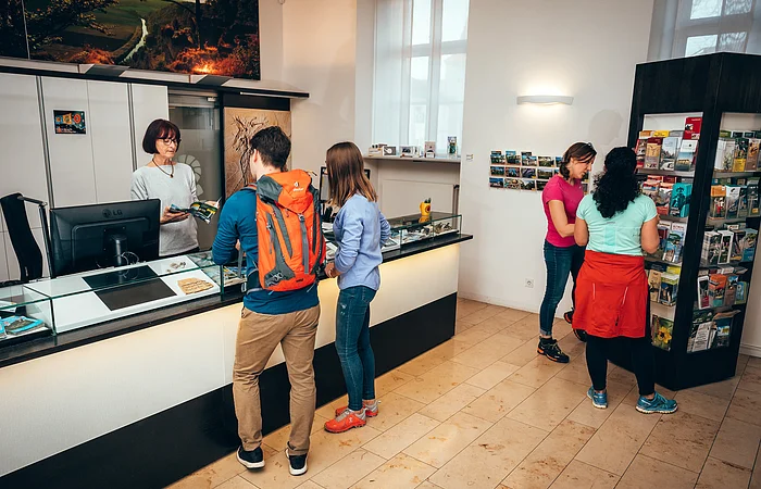 Innenraum einer Touristeninformation mit vier Personen, zwei am Tresen und zwei vor einem Prospektständer.