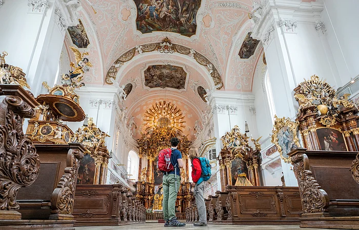Zwei Personen mit Rucksäcken stehen in einer barocken Kirche mit verzierten Holzstühlen und bemalter Decke.