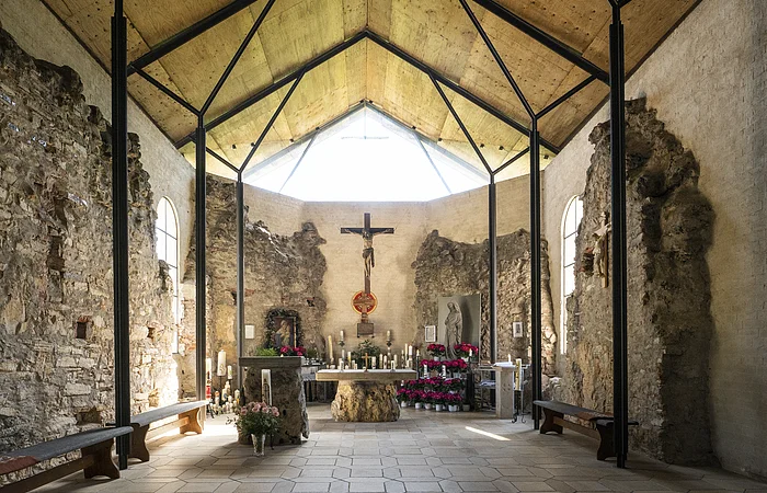 Innenraum einer Kapelle mit Altar, Kreuz, Kerzen und Blumen, umgeben von alten Steinmauern und Holzdecke