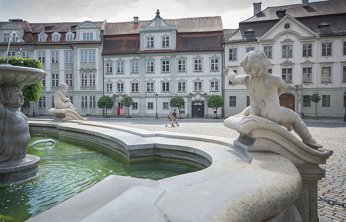Brunnen mit Puttenfiguren im Vordergrund und historische Gebäude mit roten Dächern im Hintergrund, zwei Personen gehen.