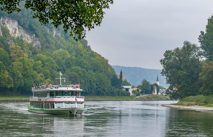 Fahrgastschiff auf Fluss vor bewaldetem Hang und Dorf mit Kirchturm bei bewölktem Himmel