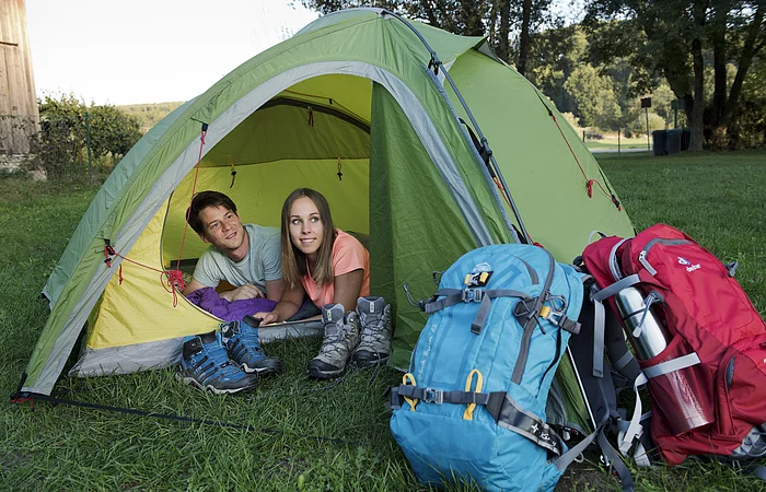 Zwei Personen liegen in einem grünen Zelt, davor Wanderschuhe und zwei große Rucksäcke auf Gras.