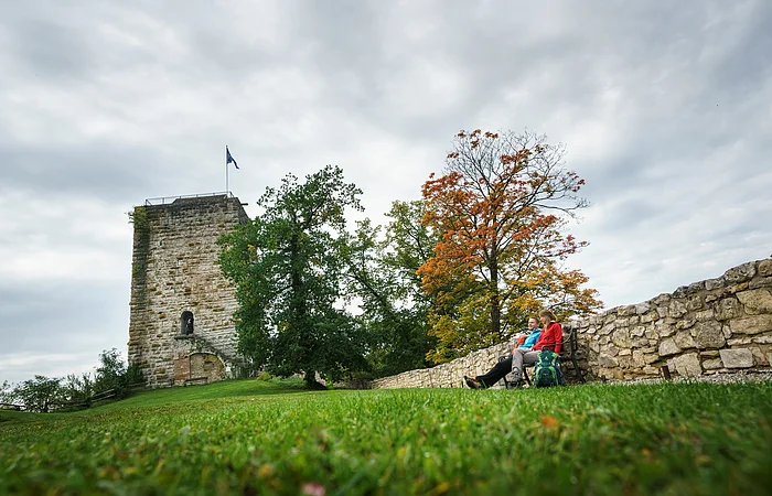 Zwei Personen sitzen auf einer Bank neben einer Steinmauer vor einem Turm und herbstlich gefärbten Bäumen.