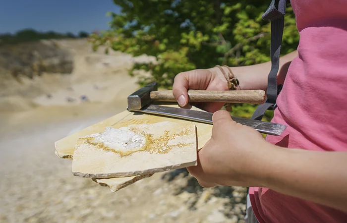 Person hält Hammer, Meißel und Gesteinsplatten mit einem weißen Mineral in einer Steinbruchumgebung