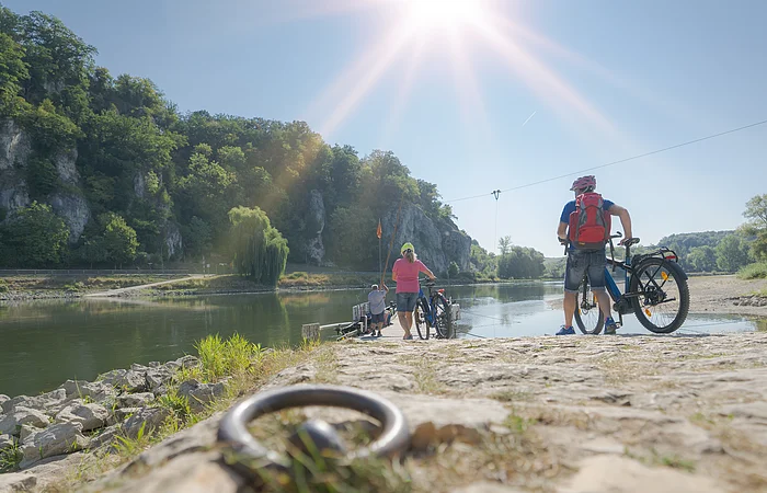 Personen mit Fahrrädern stehen an einem Flussufer mit bewaldeten Felsen im Hintergrund bei Sonnenschein.