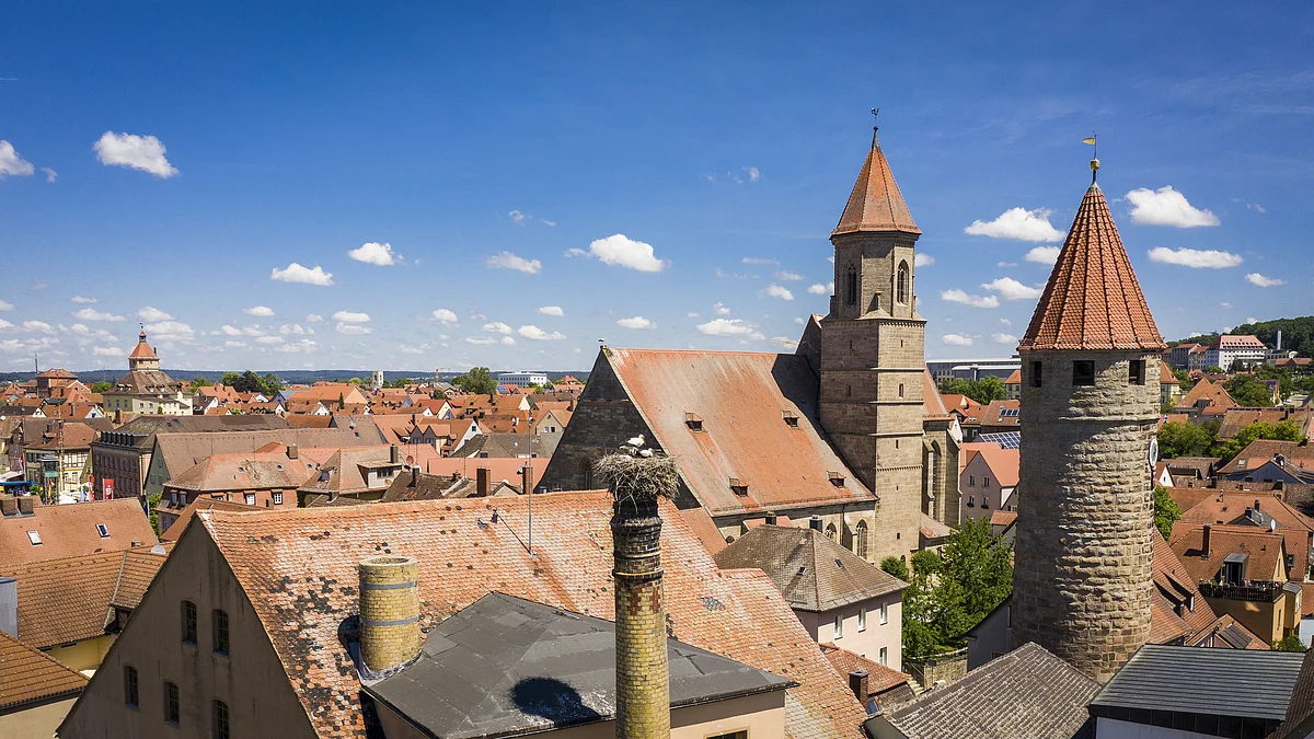 Stadtansicht mit roten Ziegeldächern, Kirchturm und rundem Steinturm unter blauem Himmel mit Wolken.
