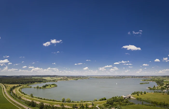 Panoramablick auf einen See mit Uferwegen, Wiesen und blauem Himmel mit vereinzelten Wolken.