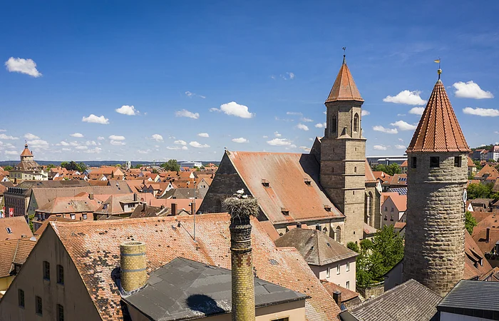 Stadtansicht mit roten Ziegeldächern, Kirchturm und rundem Steinturm unter blauem Himmel mit Wolken.