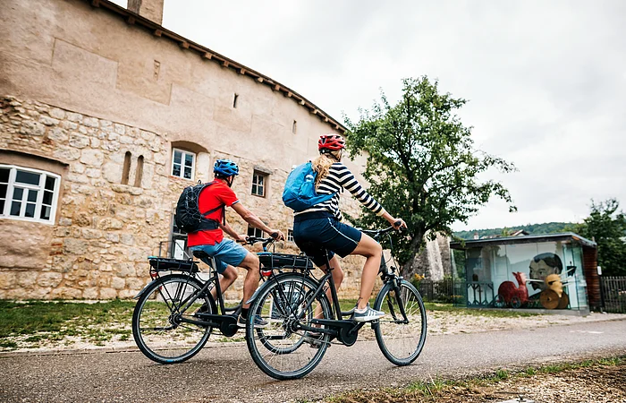 Zwei Radfahrer mit Helmen und Rucksäcken fahren an einer alten Steinmauer und einem Baum vorbei.