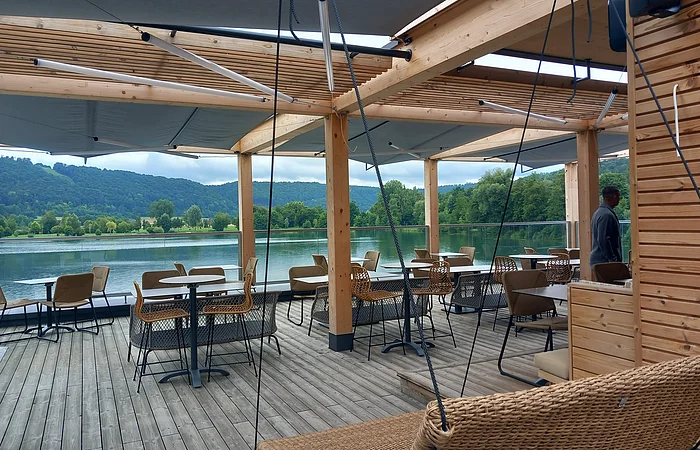 Überdachte Terrasse mit Tischen und Stühlen an einem See, im Hintergrund Bäume und bewaldete Hügel.