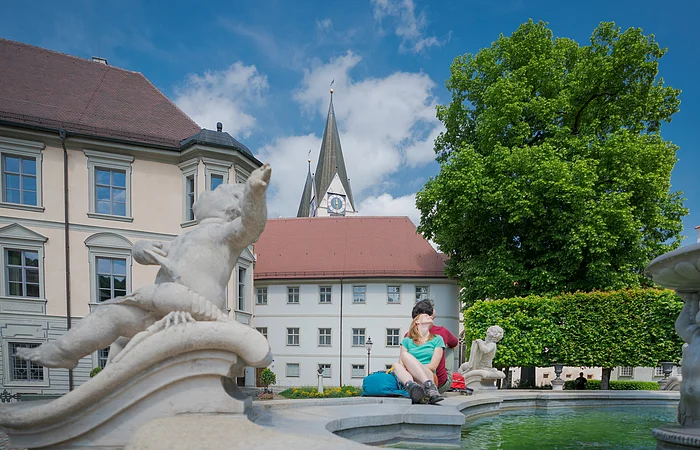 Paar sitzt am Rand eines Brunnens mit Steinfiguren vor historischen Gebäuden und einer Kirche bei blauem Himmel.
