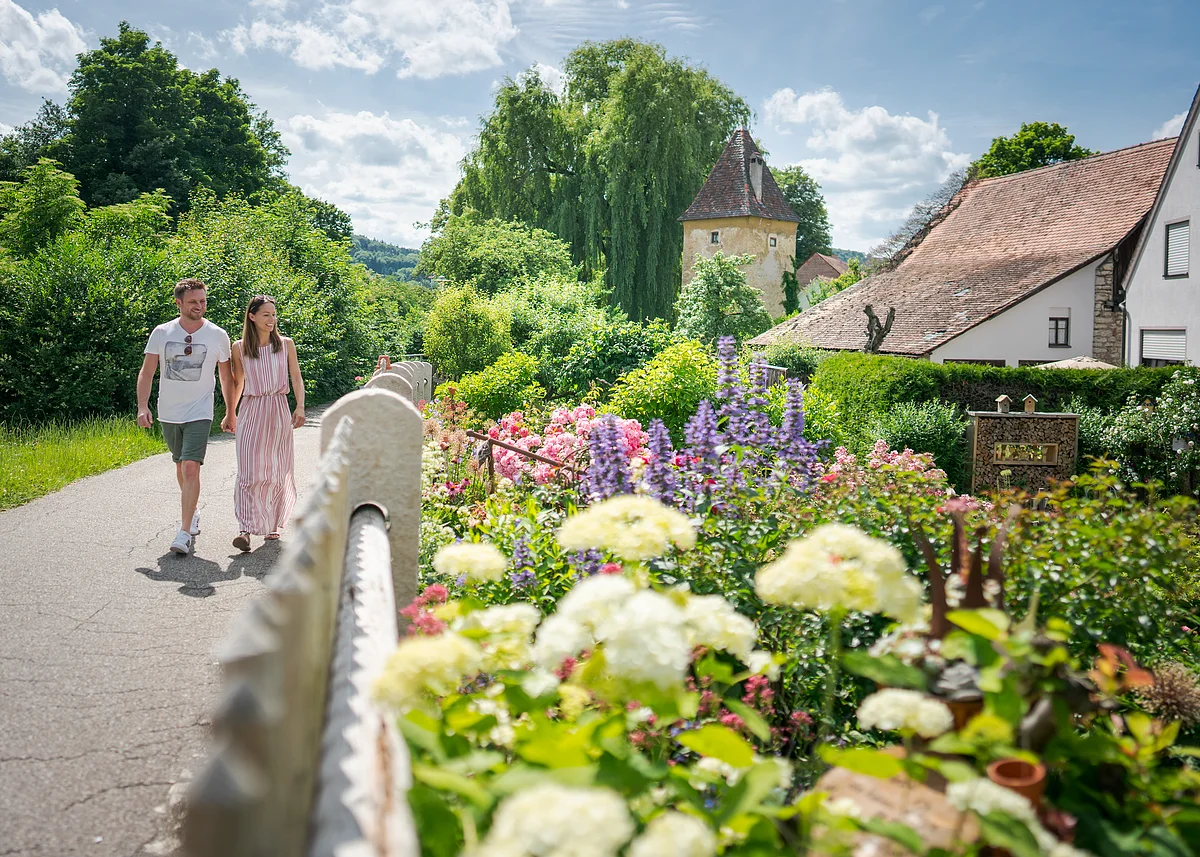Ein Mann und eine Frau – er in T-Shirt und kurzer Hose, sie im Sommerkleid – spazieren an einem Garten mit üppigen Blüten vorbei. Im Hintergrund ist ein Stadtmauerturm zu erkennen.