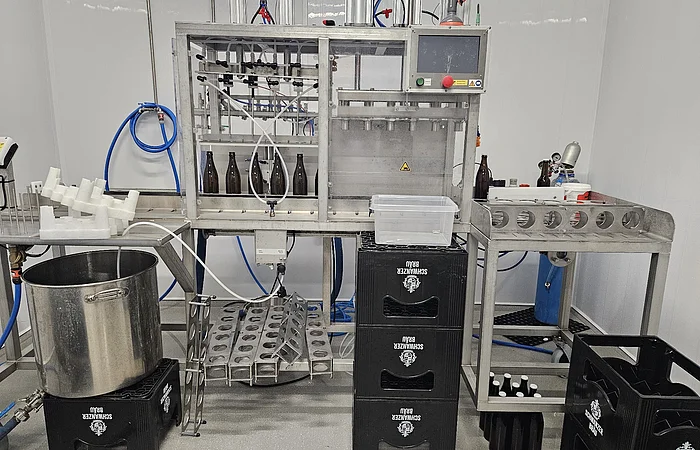 Abfüllanlage mit braunen Flaschen, Metallgestell, Kunststoffkisten und Schläuchen in einem Raum mit weißen Wänden.