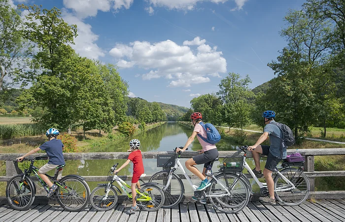 Vier Personen mit Fahrrädern auf einer Holzbrücke mit Blick auf einen Fluss und grüne Landschaft.