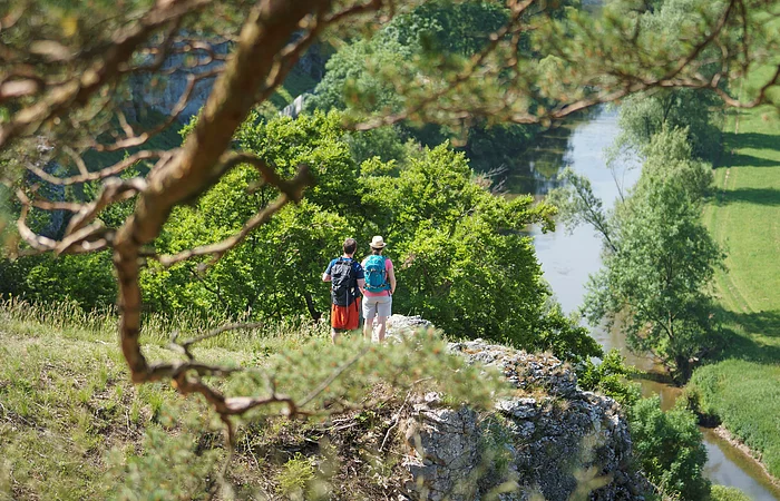 Zwei Wanderer stehen auf einem Felsen mit Blick auf einen Fluss und grüne Landschaft im Tal.