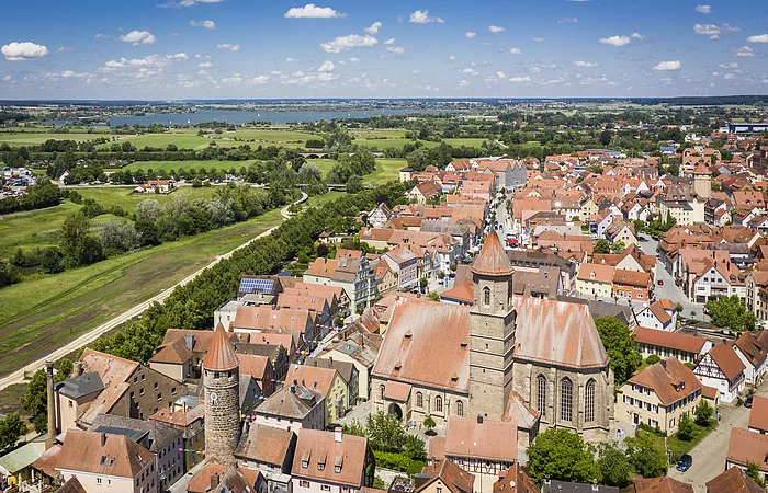 Luftaufnahme einer Stadt mit roten Dächern, zwei Türmen und einer großen Kirche, im Hintergrund grüne Felder und ein See.