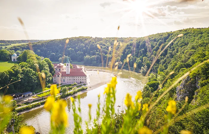 Fluss mit Schloss am Ufer, umgeben von Wald und gelben Blumen im Vordergrund bei Sonnenschein