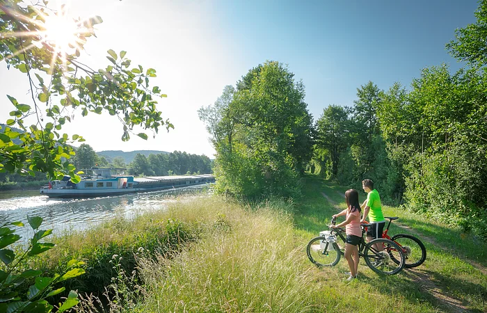 Zwei Personen mit Fahrrädern stehen an einem Flussufer und beobachten ein vorbeifahrendes Frachtschiff.