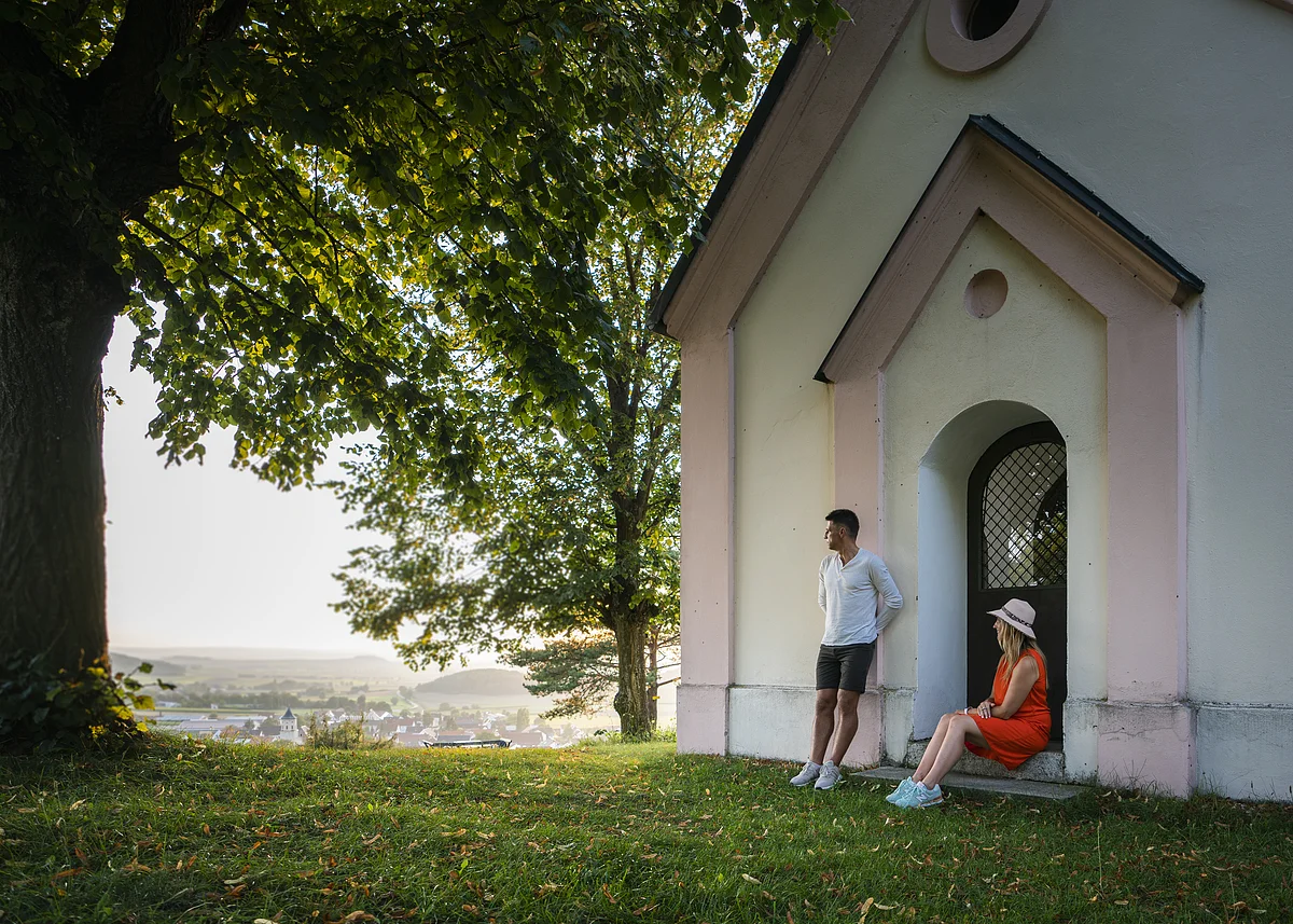 Mann lehnt an Hauswand, Frau sitzt in Türbogen, große Bäume, Dorf im Hintergrund bei Tageslicht.