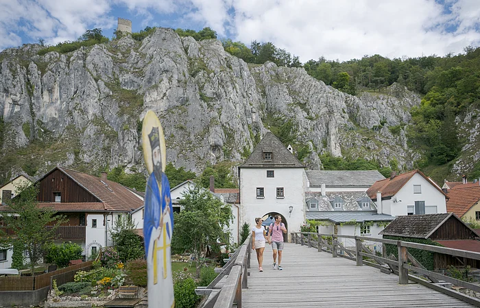 Paar geht auf einer Holzbrücke zu einem historischen Torhaus vor Felsen und Häusern unter bewölktem Himmel.