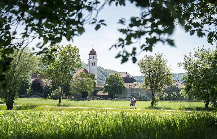 Zwei Personen gehen auf einem Weg durch eine grüne Wiese, im Hintergrund ein Kirchturm und Bäume.