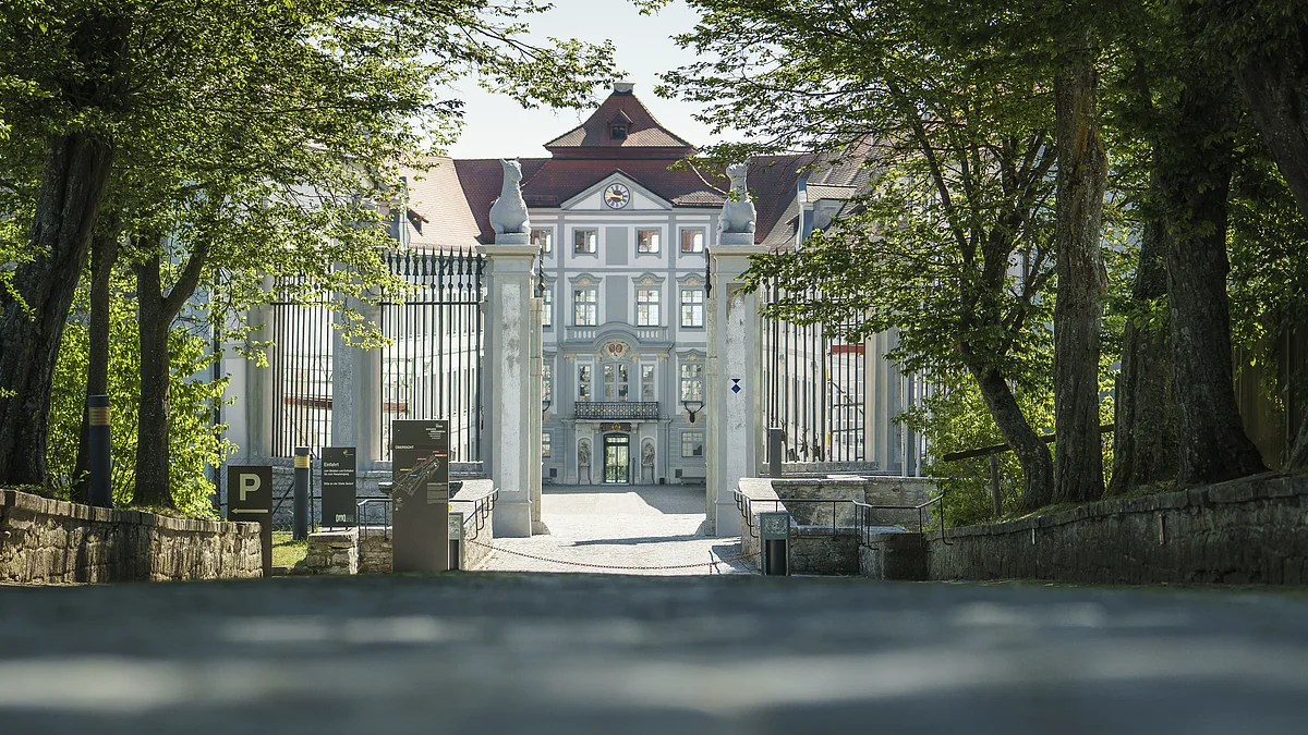 Eingangstor zu einem historischen Gebäude mit Bäumen und Schildern am Wegesrand bei Tageslicht.