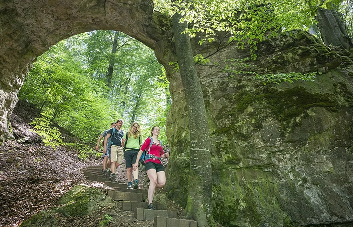 Vier Wanderer gehen einen Waldweg mit Steintreppe unter einem natürlichen Felsbogen hinab.