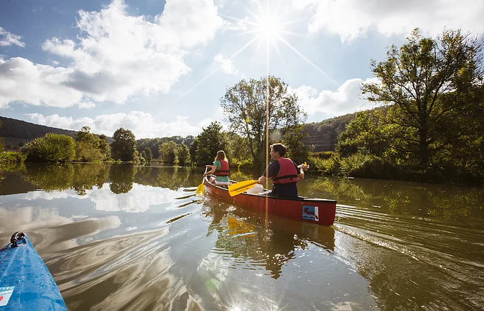 Zwei Personen paddeln in einem roten Kanu auf einem ruhigen Fluss bei sonnigem Himmel mit Wolken.