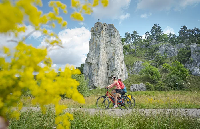 Zwei Radfahrer fahren auf einem Weg vor einem hohen Felsen mit Kreuz, gelbe Blumen im Vordergrund, bewölkter Himmel