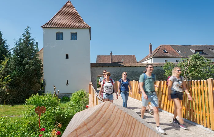Fünf Personen mit Rucksäcken gehen auf einem gepflasterten Weg neben einem weißen Turm mit rotem Dach bei klarem Himmel.