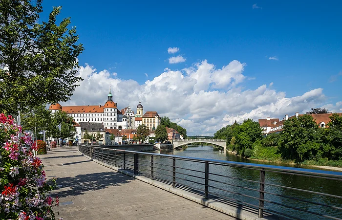 Fluss mit Brücke, rechts Bäume und Häuser, links Blumen, Baum und Schloss im Hintergrund bei blauem Himmel.
