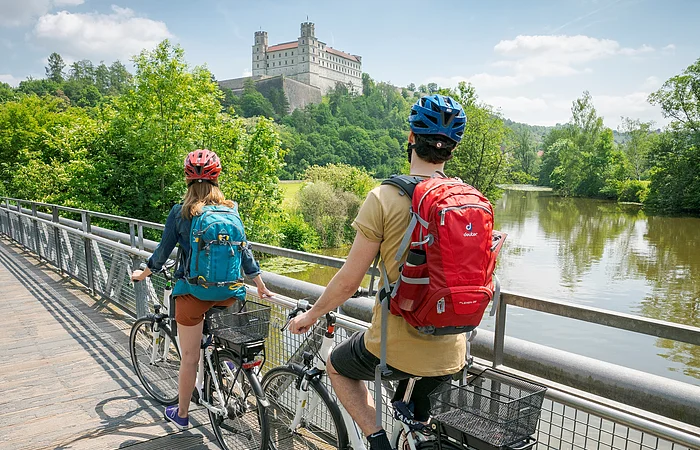 Zwei Radfahrer mit Rucksäcken und Helmen stehen auf einer Holzbrücke und blicken auf ein Schloss auf einem bewaldeten Hügel.