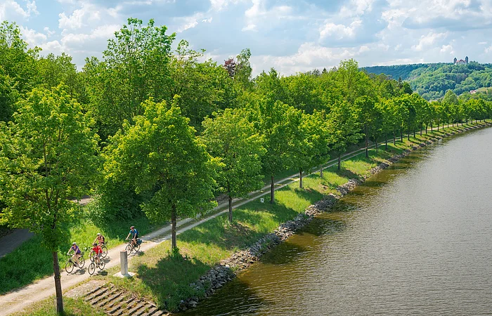 Fünf Radfahrer auf einem Weg entlang eines Flusses mit Bäumen und Hügeln im Hintergrund bei bewölktem Himmel.
