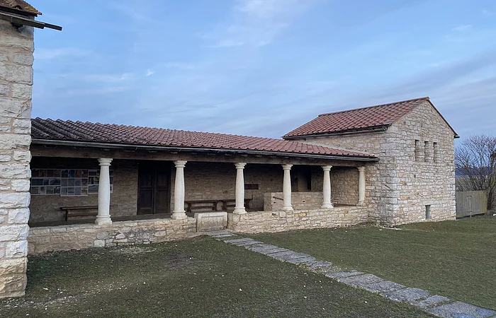 Steinernes Gebäude mit Säulen und rotem Ziegeldach vor bewölktem Himmel und Rasenfläche