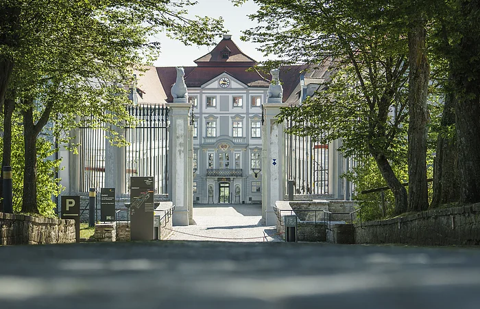 Eingangstor zu einem historischen Gebäude mit Bäumen und Schildern am Wegesrand bei Tageslicht.