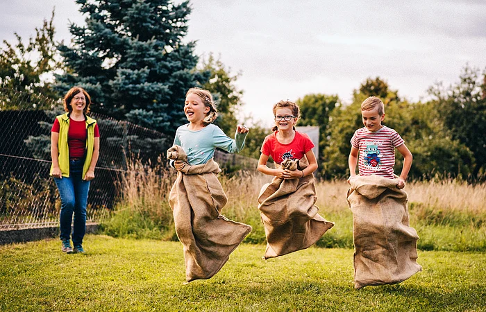 Drei Kinder springen in Jutesäcken auf einer Wiese, eine Frau beobachtet sie lachend im Hintergrund.