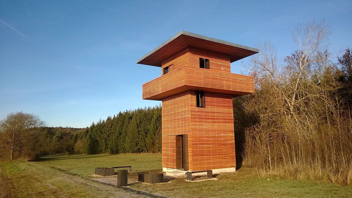 Holzbeobachtungsturm mit umlaufendem Balkon in einer Wiesenlandschaft neben Wald und kahlen Bäumen bei blauem Himmel